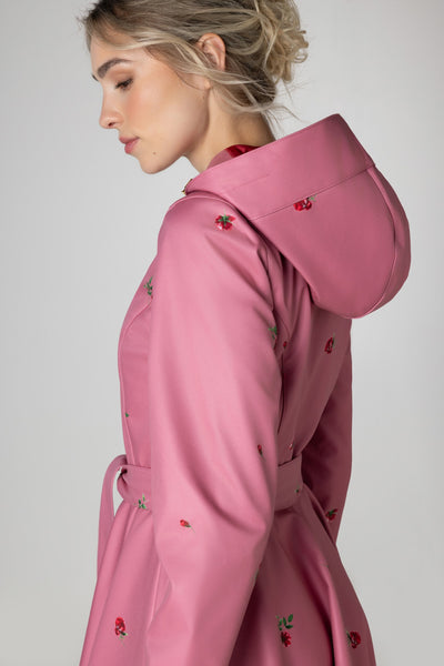 A woman in profile wearing RainSisters pink hooded raincoat Raspberry Sorbet with a tie belt and red floral print, photographed against a light gray studio background.