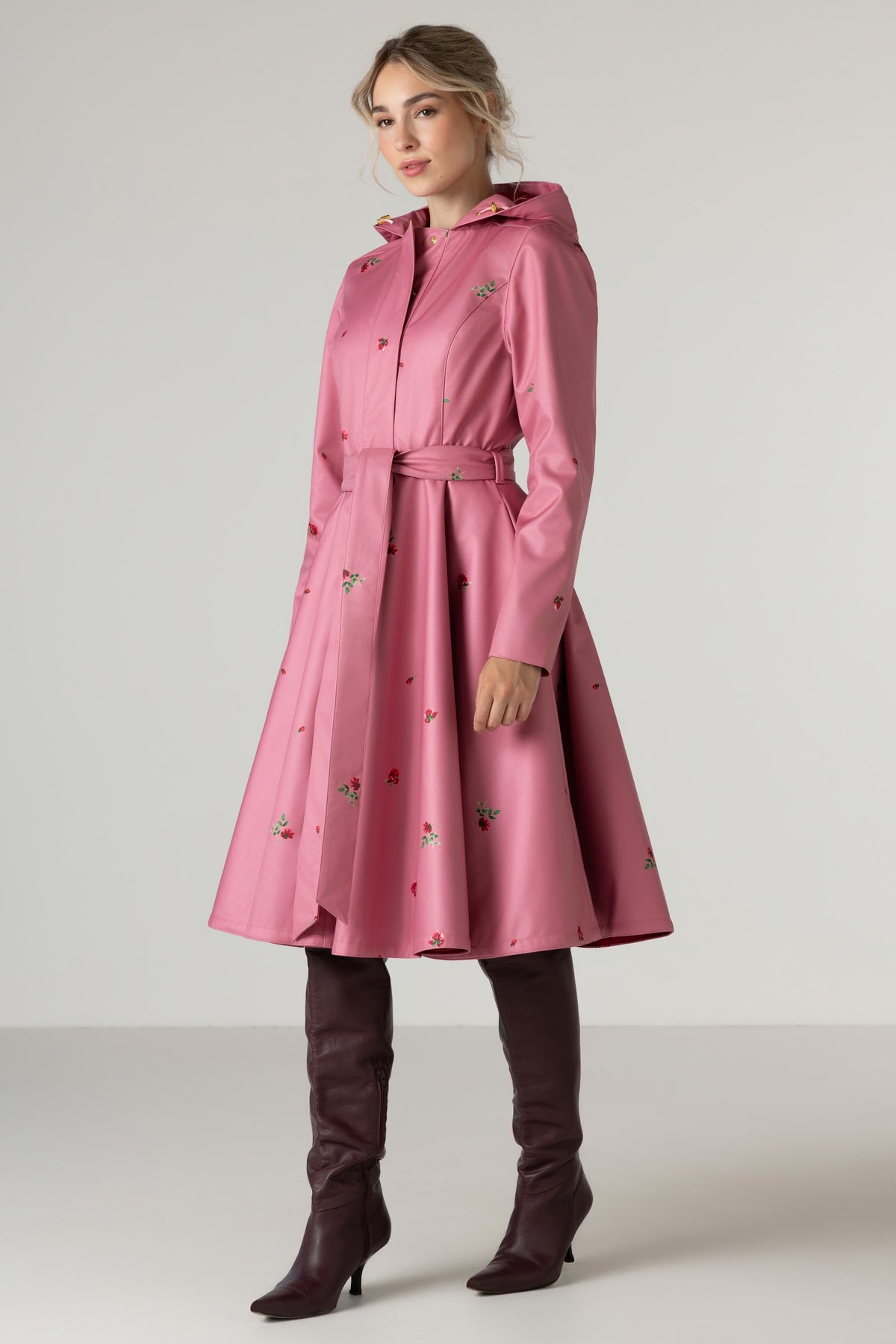A woman models RainSisters pink hooded raincoat Raspberry Sorbet with a matching waist belt and small floral print, paired with dark brown knee-high boots, against a neutral studio background.