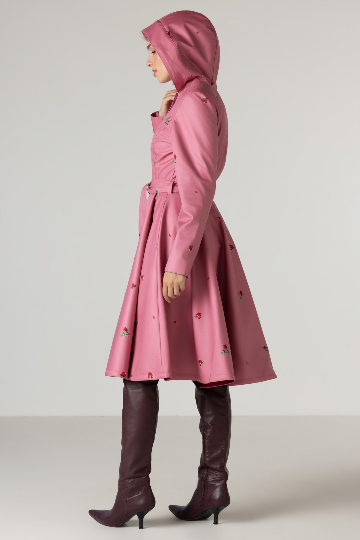 A model is shown in profile wearing RainSisters pink hooded raincoat Raspberry Sorbet with small red rose print, paired with dark brown knee-high leather boots, against a neutral light gray studio background.