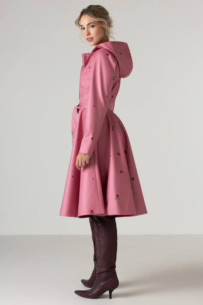 A woman modeling RainSisters pink hooded raincoat Raspberry Sorbet with a belted waist and small red and green rose flower print, paired with dark brown knee-high boots, against a plain light gray studio background.