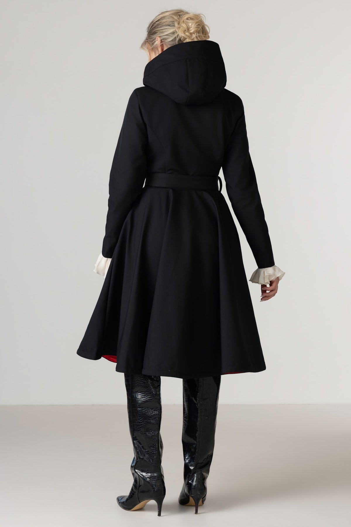 A woman shown from the back wearing RainSisters long black hooded raincoat Raven Red with a waist tie and flared skirt, paired with black knee-high boots and white gloves, against a plain light gray studio background.