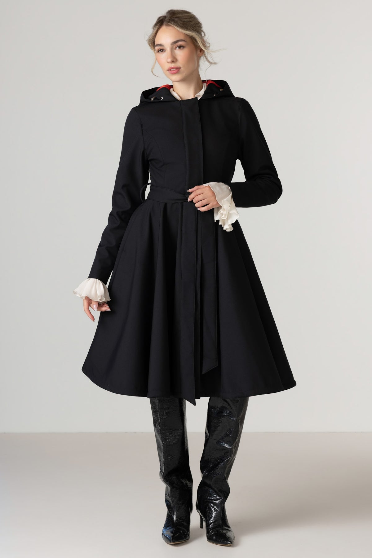 A woman models RainSisters black hooded coat Raven Red with a matching waist tie, styled with white ruffled cuffs, black leather knee-high boots, and a white collared shirt underneath, set against a plain light gray studio background.