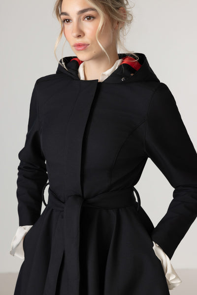 A woman wearing RainSisters black hooded waterproof coat Raven Red with a tie waist belt, layered over a cream shirt, photographed against a light gray studio background.