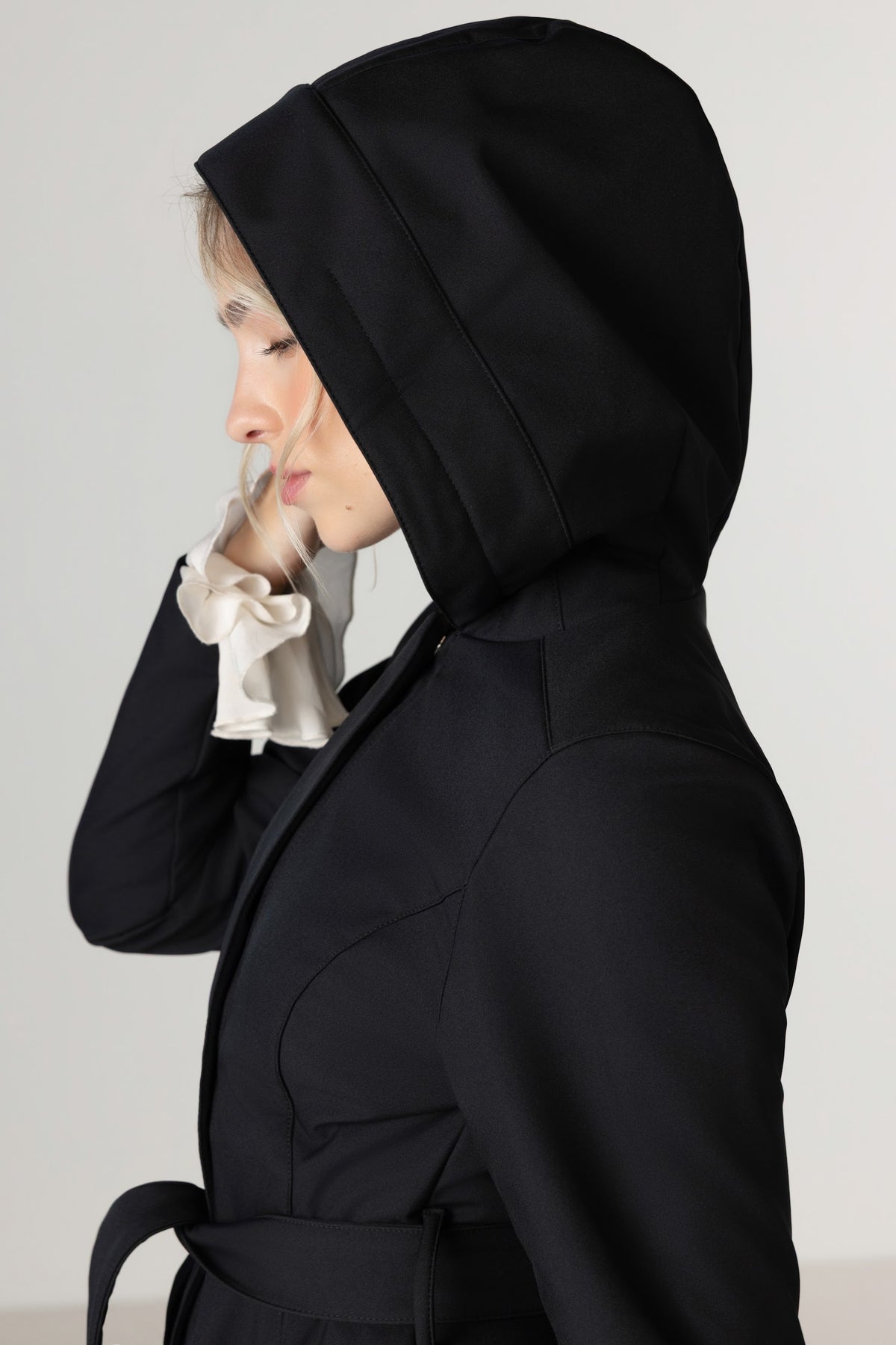 A woman wearing RainSisters black hooded raincoat with a matching waist tie, shown in profile against a light gray studio background.