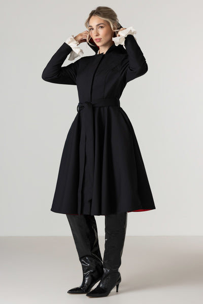 A woman models RainSisters black long-sleeve A-line waterproof dress coat Raven Red with a matching waist tie belt, styled with contrasting white ruffled cuffs. She is wearing black pointed-toe heeled boots against a neutral light gray studio background.