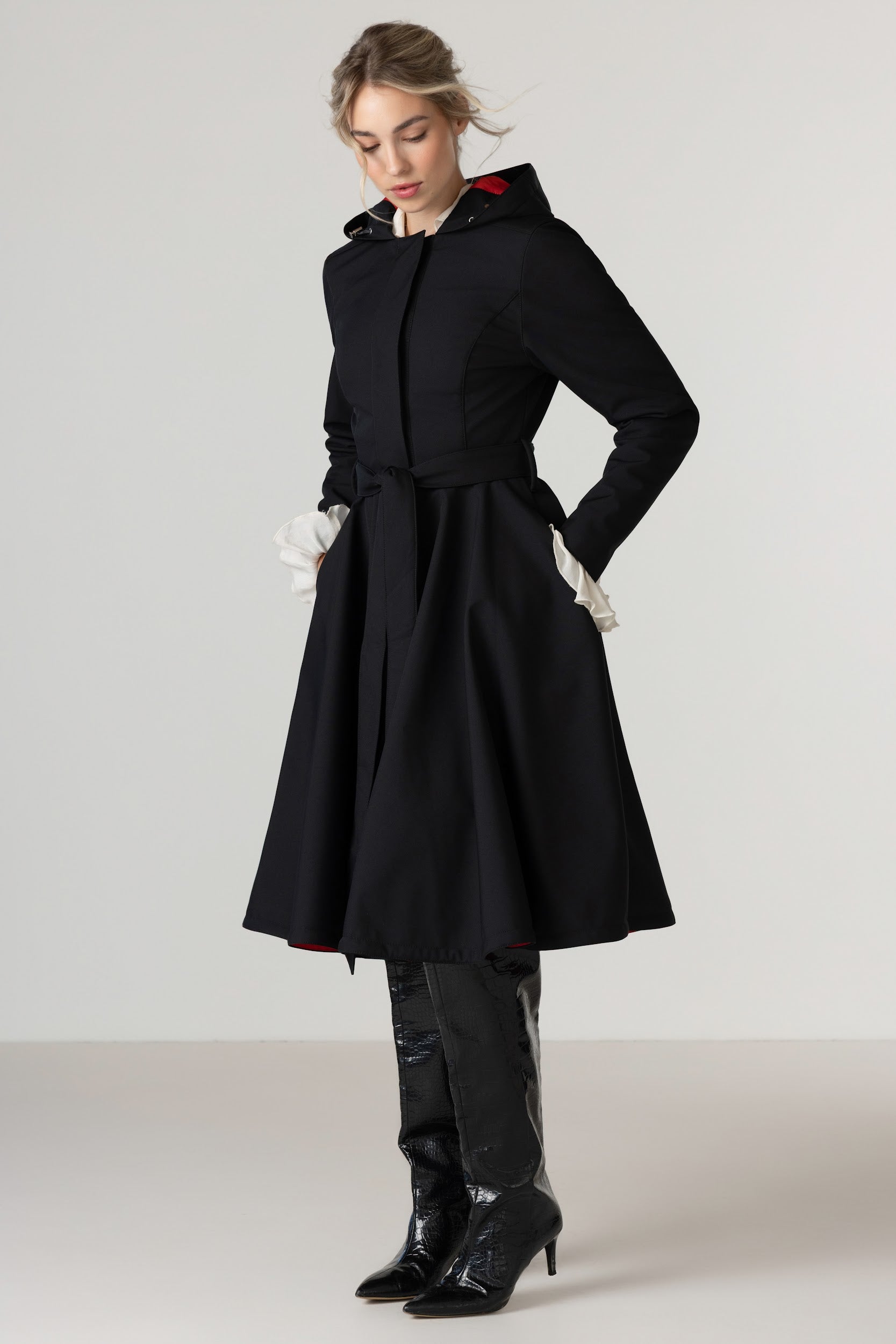A woman modeling RainSisters black hooded raincoat Raven Red with a belted waist and black knee-high boots against a plain light gray studio background.