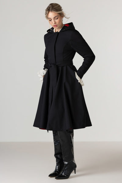 A woman modeling RainSisters black hooded raincoat Raven Red with a belted waist and black knee-high boots against a plain light gray studio background.
