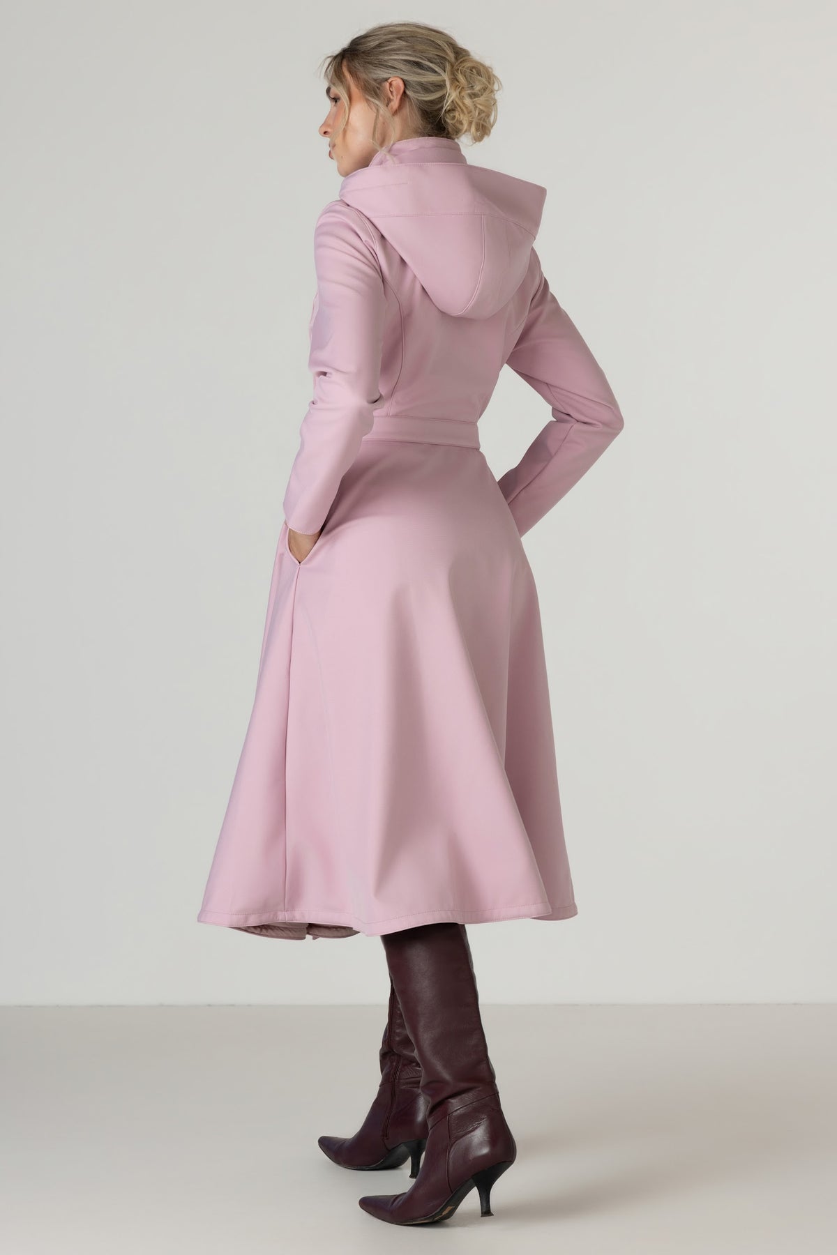 A woman shown from the back wearing RainSisters light pink hooded long coat Rosie Bow with a waist tie and dark brown heeled boots, photographed against a plain light gray studio background.