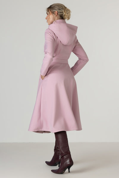 A woman shown from the back wearing RainSisters light pink hooded long coat Rosie Bow with a waist tie and dark brown heeled boots, photographed against a plain light gray studio background.