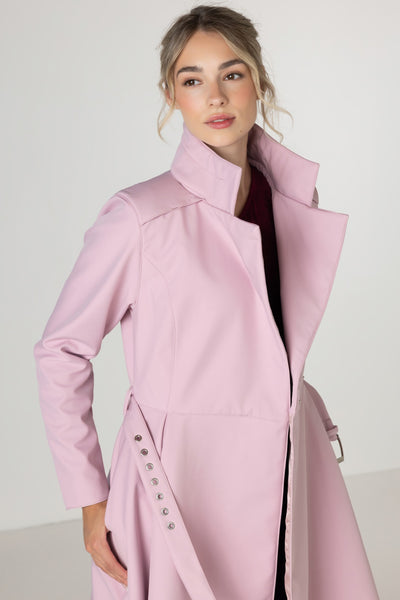A woman wearing RainSisters light pink mid-length trench coat Rosie Bow with a high collar and silver-tone decorative buttons on the side, photographed against a plain light gray studio background.