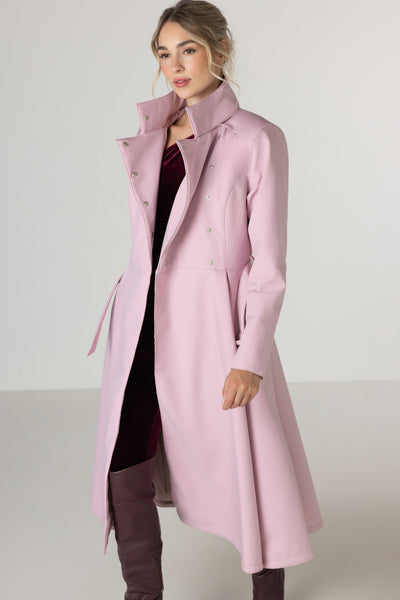 A woman modeling RainSisters midseason long, light pink double-breasted coat Rosie Bow with a high collar, worn over a dark burgundy top and brown over-the-knee boots, set against a plain light gray studio background.