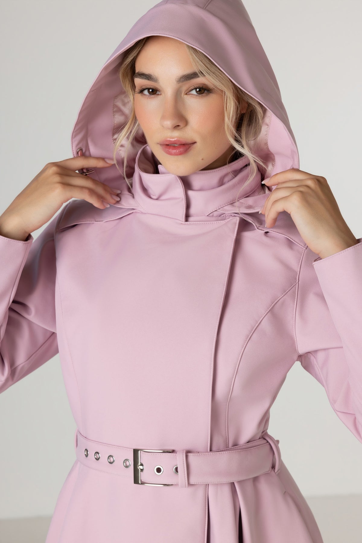 A woman wearing RainSisters light pink hooded raincoat Rosie Bow with a matching belt and silver buckle, posing with her hands on the hood.