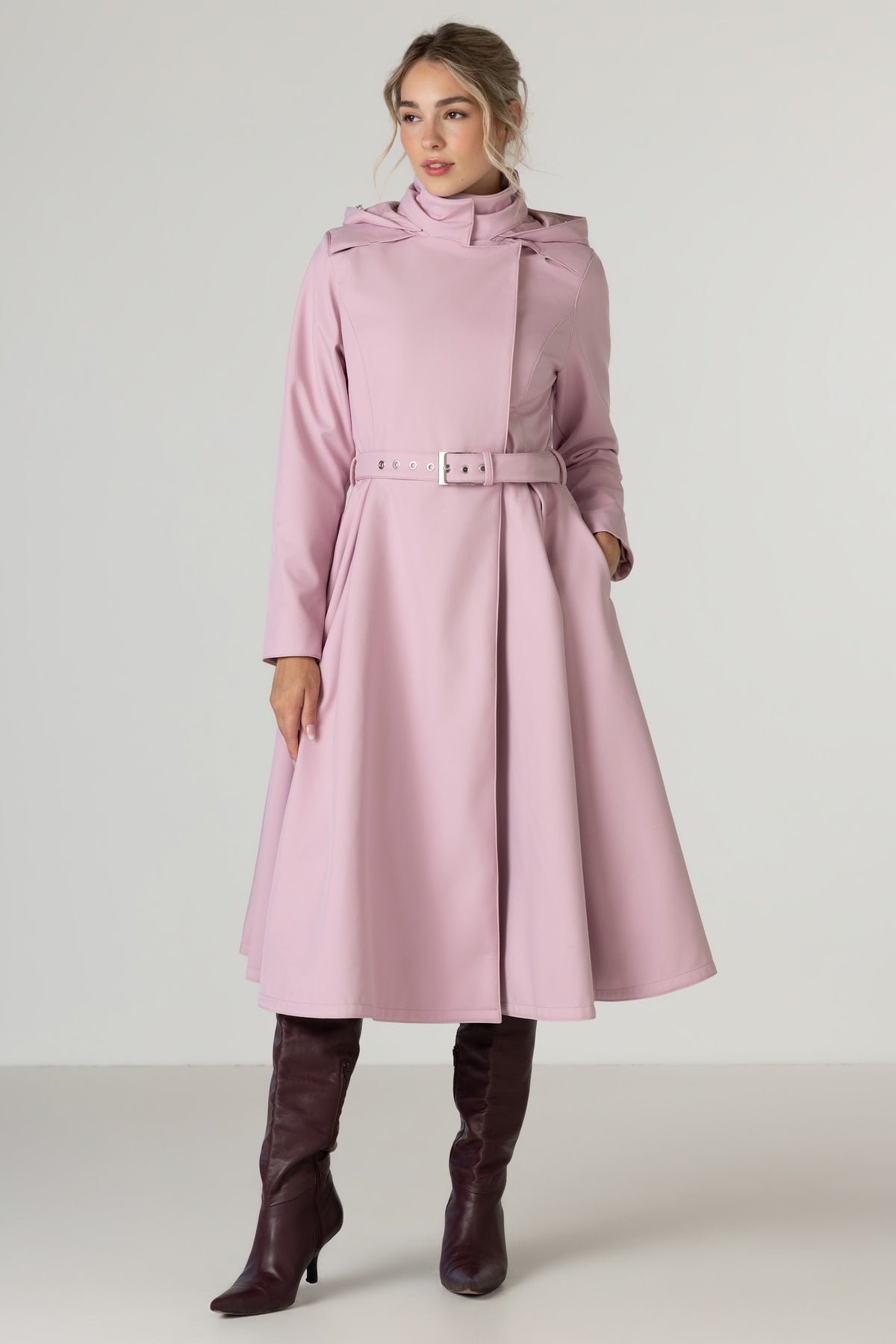A woman modeling RainSisters waterproof coat Rosie Bow - a dusty pink belted coat with a high structured collar and wide lapels, paired with dark brown knee-high boots, against a light gray studio background.