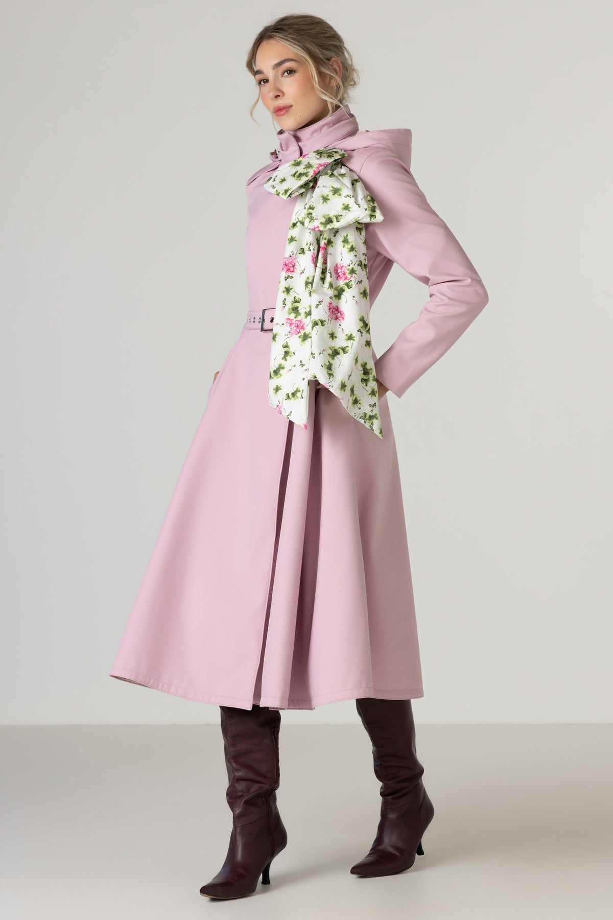 A model wearing a light pink waterproof trench coat by RainSisters with a matching belt, a detachable white scarf with a pink and green floral print, and dark brown knee-high leather boots, posed against a neutral light gray studio background.