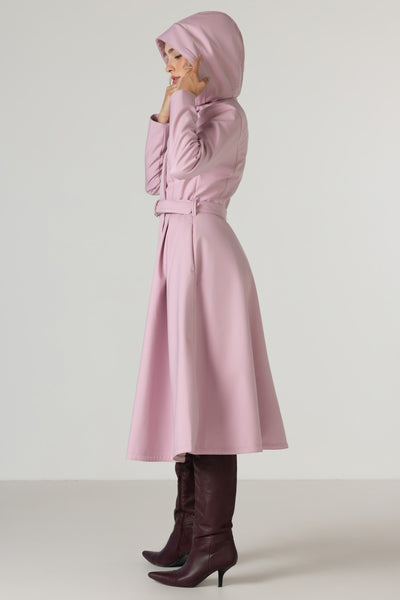 A woman wearing RainSisters light pink hooded trench coat Rosie Bow with a matching belt, styled with dark brown knee-high leather boots, shown in profile against a neutral studio background.