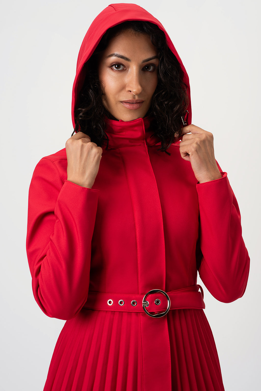 A woman wearing RainSisters bright red hooded coat Scarlet Waltz with a high collar and a matching belt with a silver buckle, posing against a light gray studio background.