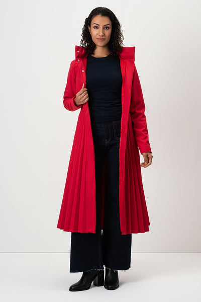 A woman modeling RainSisters long, bright red pleated trench coat Scarlet Waltz over a black top and dark wide-leg pants, standing against a plain light gray studio background.