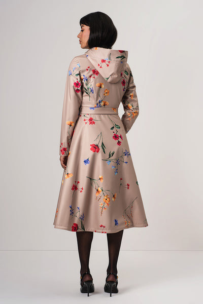 A woman viewed from the back wearing RainSisters beige hooded coat Spring Bloom with a colorful floral print, paired with black tights and black high heels, against a plain light gray studio background.