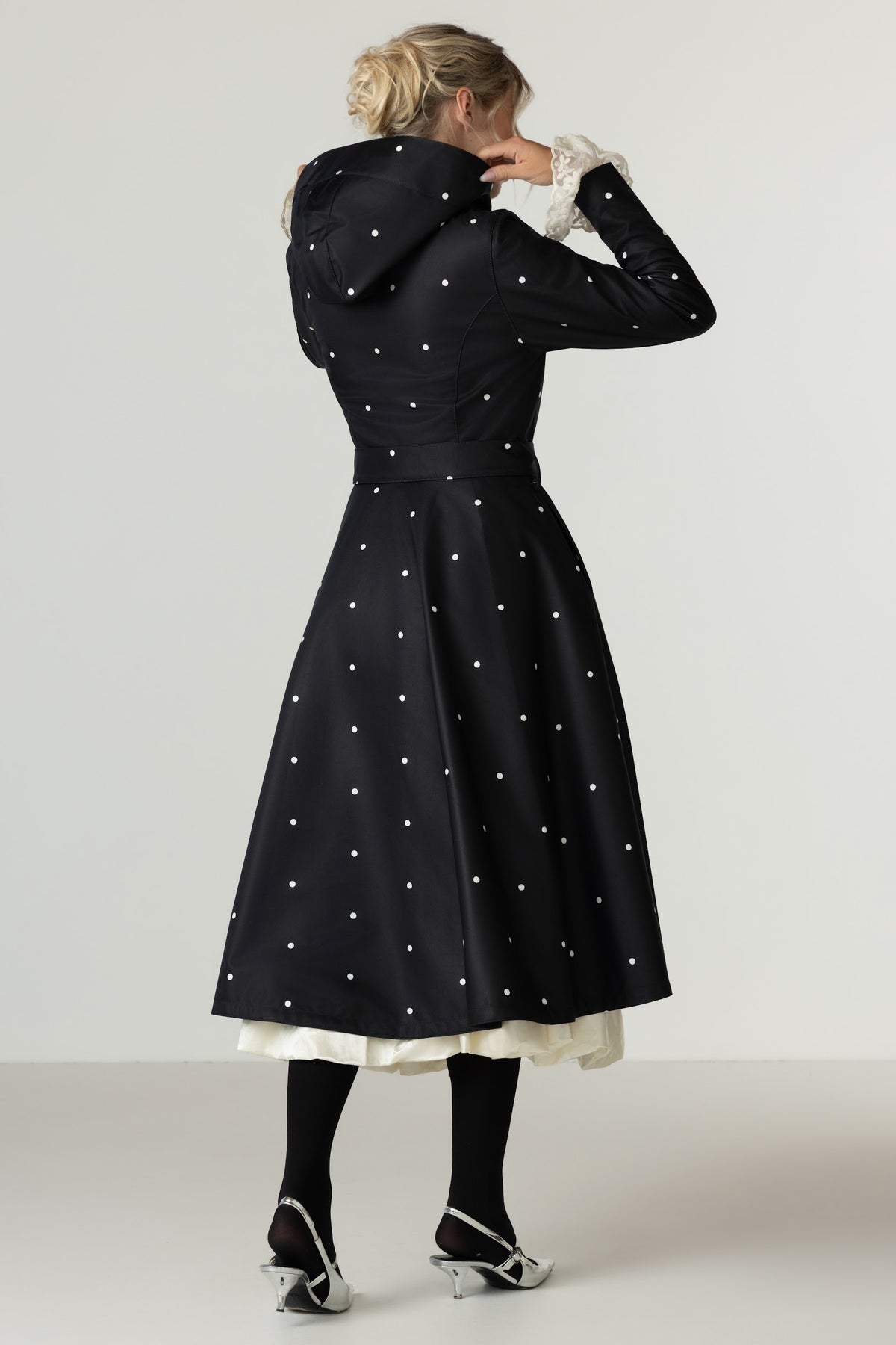 A woman photographed from the back wearing RainSisters black longline coat Timeless Polka Dots with white polka dots, styled with black tights, and silver heels against a light gray studio background.