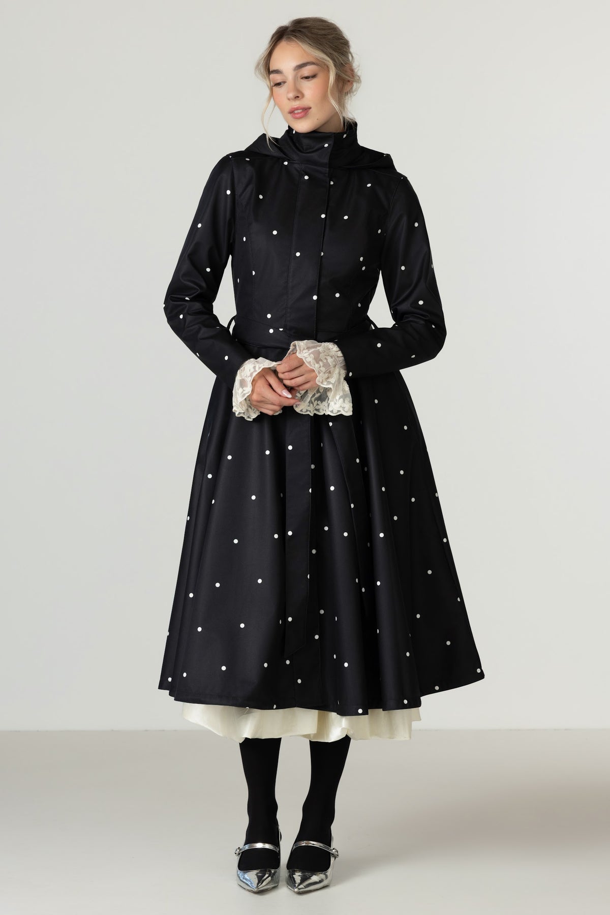 A woman models RainSisters black A-line waterproof coat Timeless Polka Dots with white polka dots, featuring a high collar and detachable hood. She is wearing black tights and silver shoes against a plain light gray studio background.