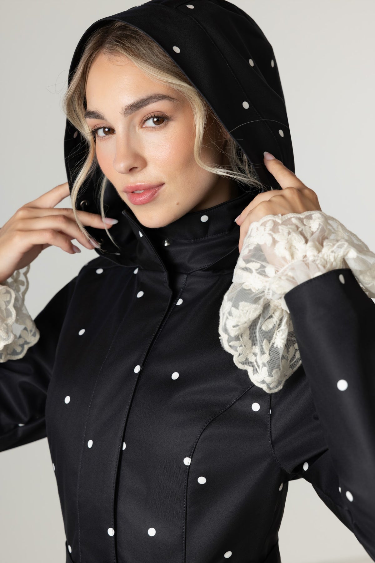 A woman wearing RainSisters black hooded coat Timeless Polka Dots with white polka dots wtyled with lace ruffled cuffs, posing against a light gray studio background.