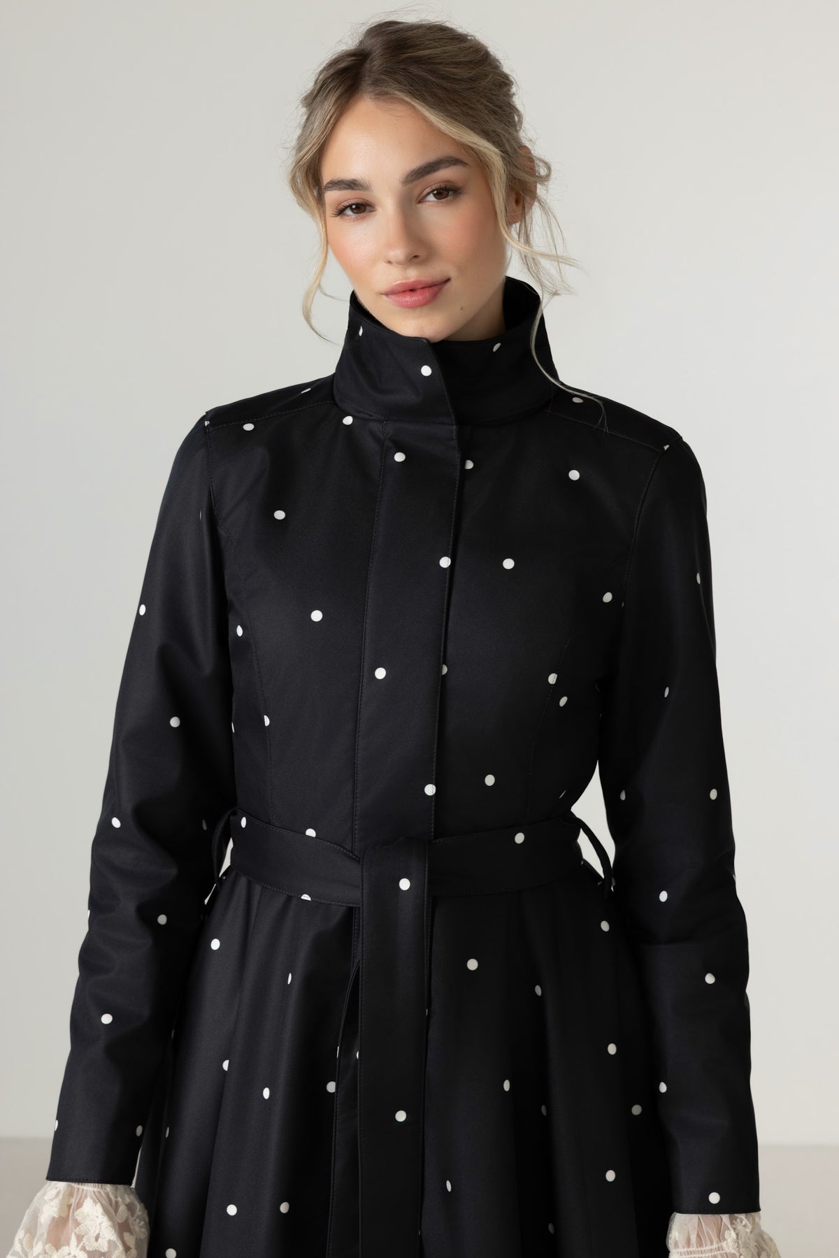 A woman wearing RainSisters black long-sleeve coat Timeless Polka Dots with white polka dots and a matching waist tie, photographed against a light gray studio background.