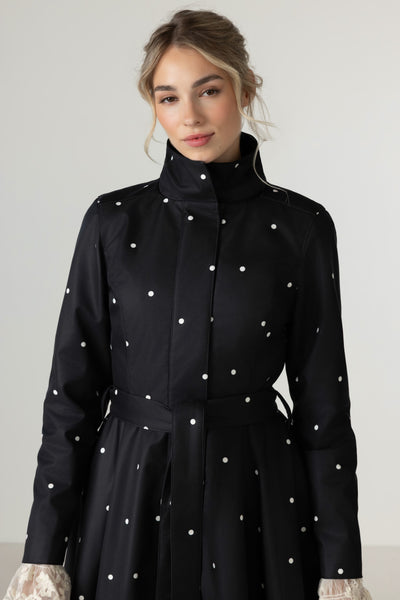 A woman wearing RainSisters black long-sleeve coat Timeless Polka Dots with white polka dots and a matching waist tie, photographed against a light gray studio background.