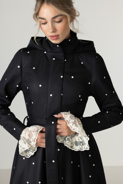A woman wearing RainSisters black hooded raincoat Timeless Polka Dotswith white polka dots and detachable hood, photographed against a plain light gray studio background.