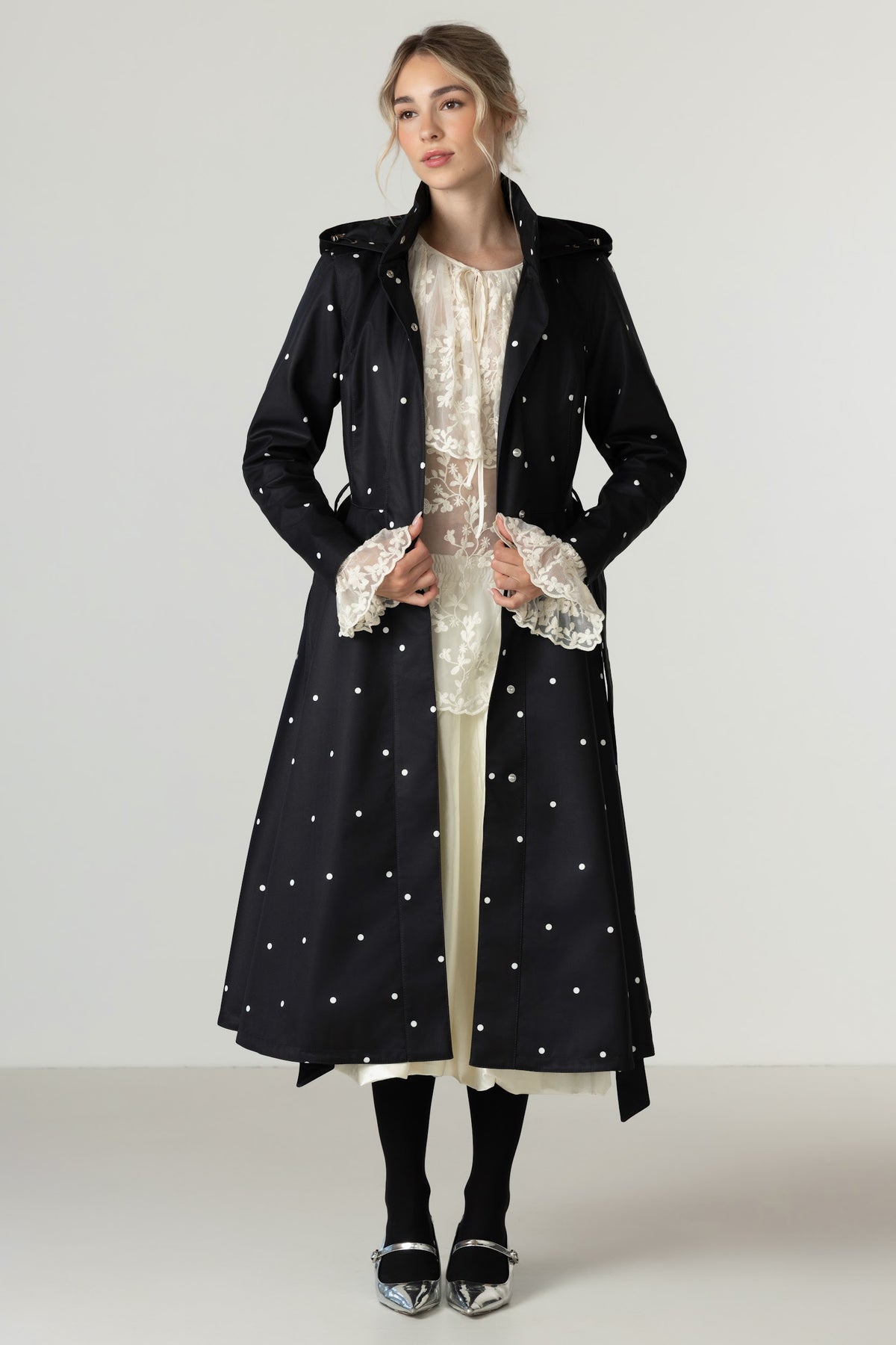 A model stands against a light gray studio background wearing RainSisters long black coat Timeless Polka Dots with white polka dots over a cream lace-trimmed dress, black tights, and silver shoes.