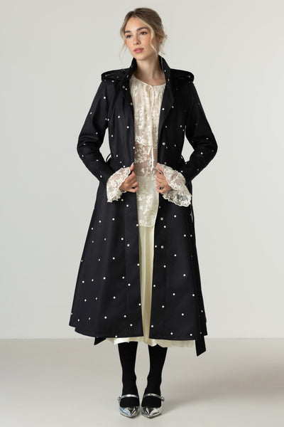 A model stands against a light gray studio background wearing RainSisters long black coat Timeless Polka Dots with white polka dots over a cream lace-trimmed dress, black tights, and silver shoes.