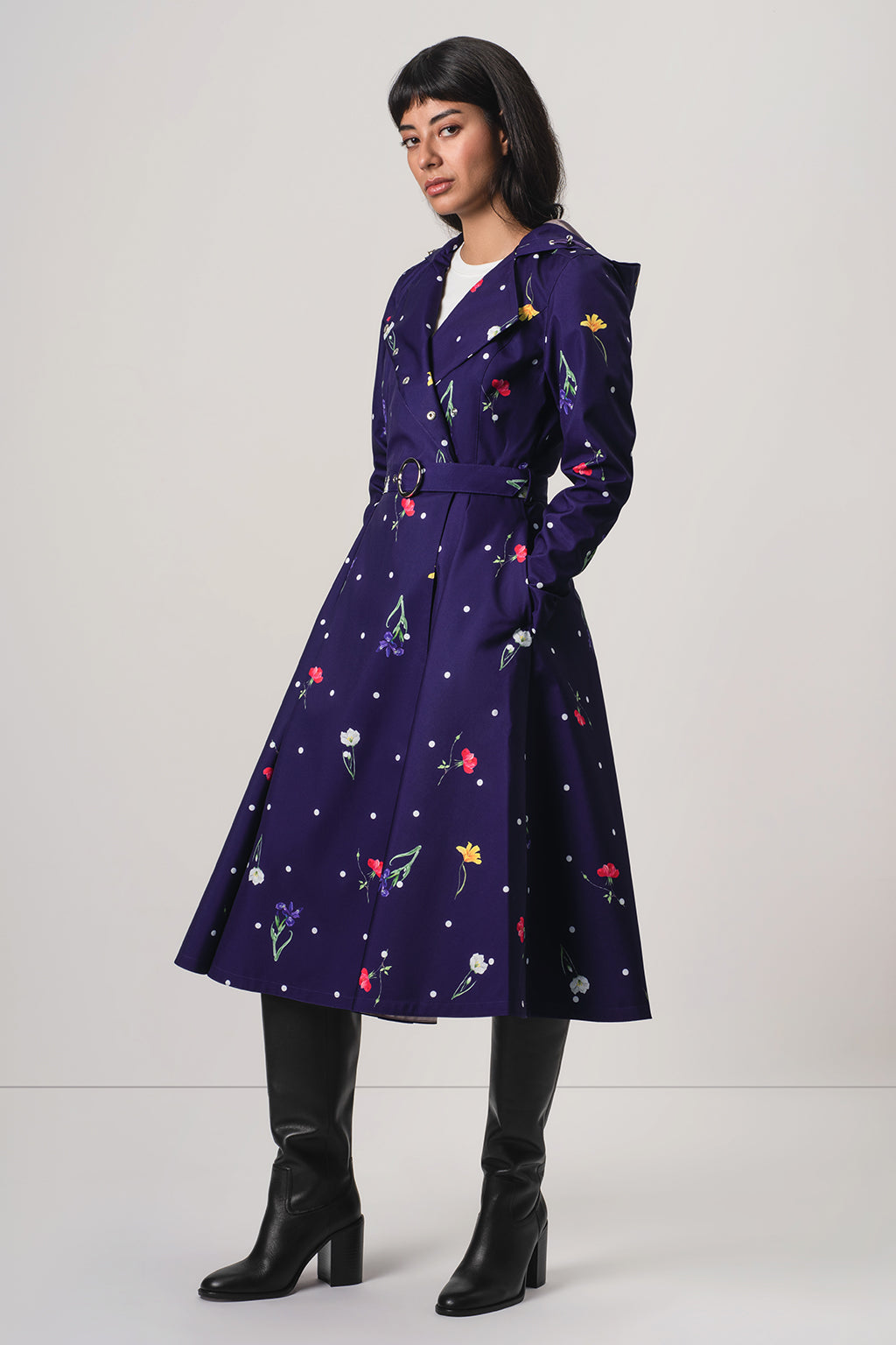 A model wearing RainSisters dark purple A-line trench coat Violet Bloom with a colorful floral and star pattern, styled with black knee-high boots against a light gray studio background.