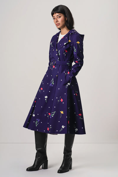 A model wearing RainSisters dark purple A-line trench coat Violet Bloom with a colorful floral and star pattern, styled with black knee-high boots against a light gray studio background.