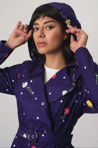 A woman wearing RainSisters dark purple hooded raincoat Violet Bloom with white polka dots and colorful floral print, posing against a light gray studio background.