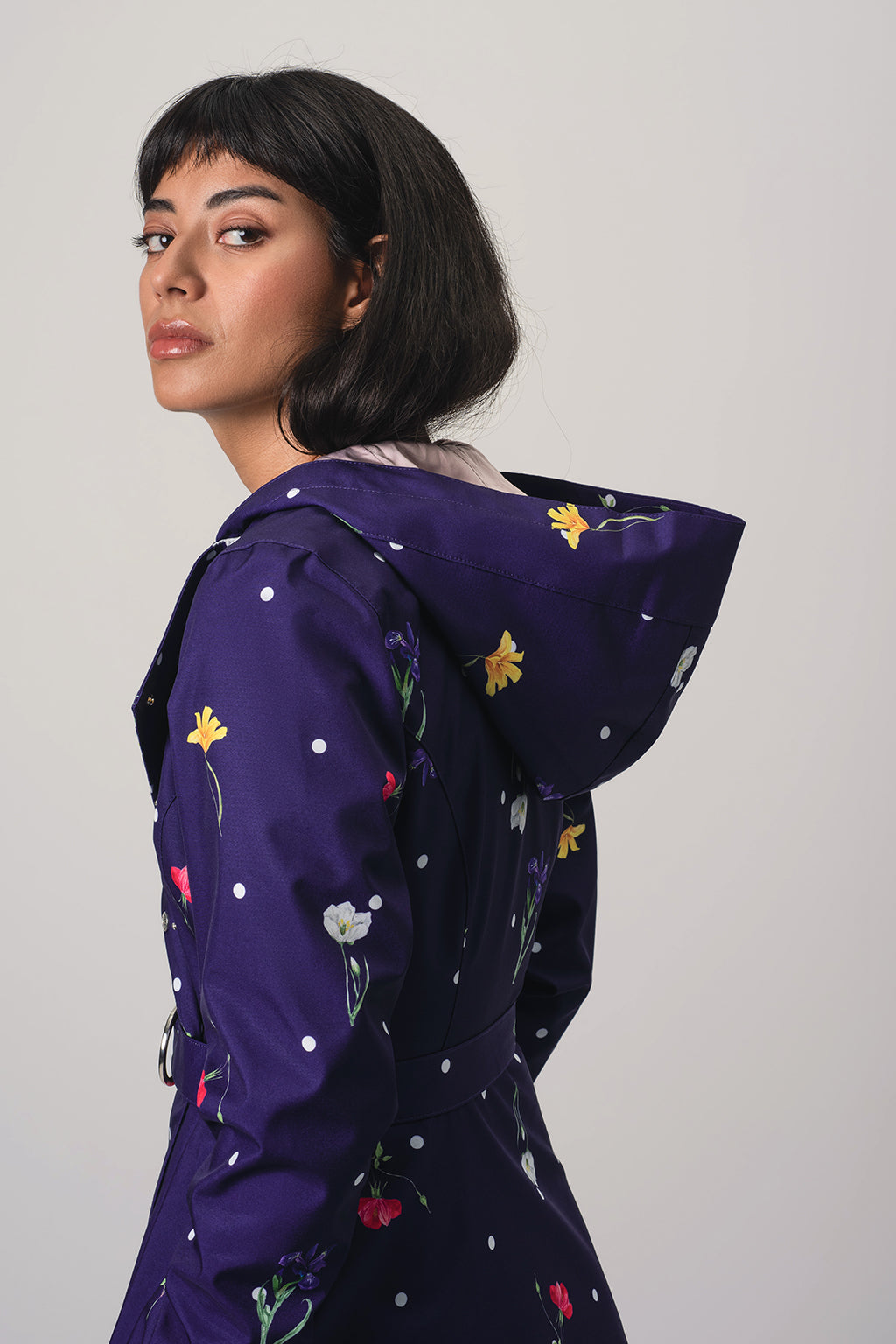 A woman wearing RainSisters dark purple hooded raincoat Violet Bloom with a colorful floral print and white polka dots, photographed against a plain light gray studio background.