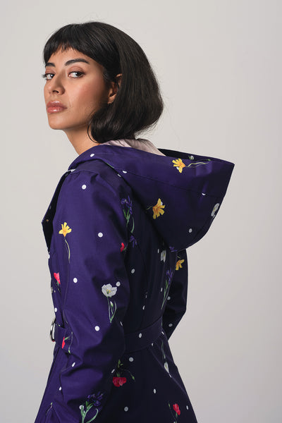 A woman wearing RainSisters dark purple hooded raincoat Violet Bloom with a colorful floral print and white polka dots, photographed against a plain light gray studio background.