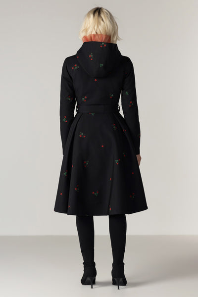 A model viewed from the back wearing RainSisters black hooded raincoat Wild Strawberry with a small red and green floral pattern, paired with black tights and black ankle boots against a light gray studio background.