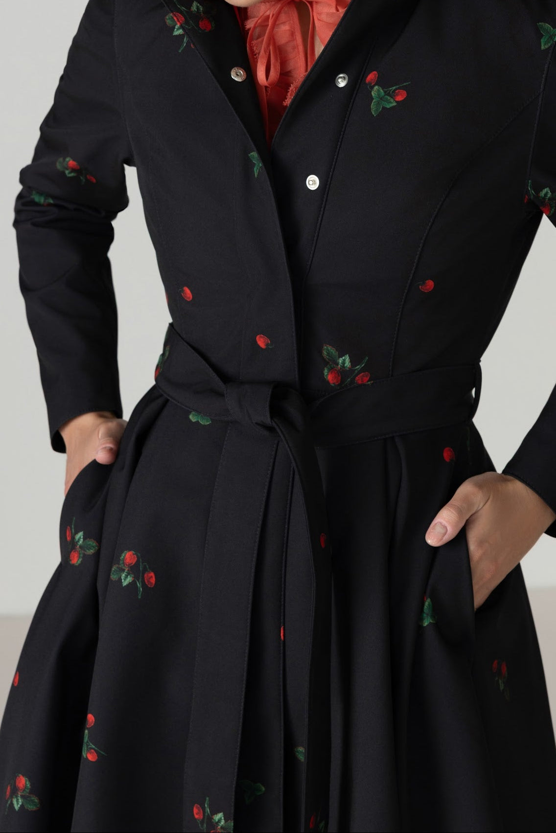 A blonde model wearing RainSisters black long-sleeve belted raincoat with red and green strawberry print, layered over a rust-orange collared shirt, against a plain light gray studio background.
