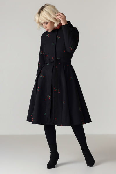 A woman modeling RainSisters black A-line dress coat Wild Strawberry with a red and green strawberry pattern, styled with black tights and black ankle boots against a light gray studio background.