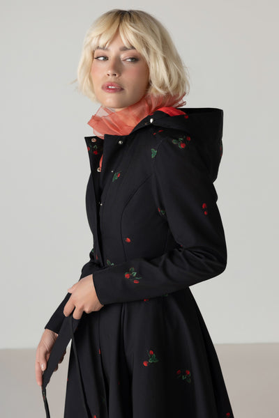 A blonde model wearing RainSisters black longline waterproof hooded coat Wild Strawberry with red and green strawberry print, styled with a sheer coral top, against a neutral light gray background.