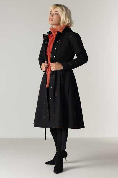 A model wearing RainSisters long black coat Wild Strawberry with small colorful strawberry print, layered over a rust-orange scarf and black tights, standing against a neutral light gray studio background.