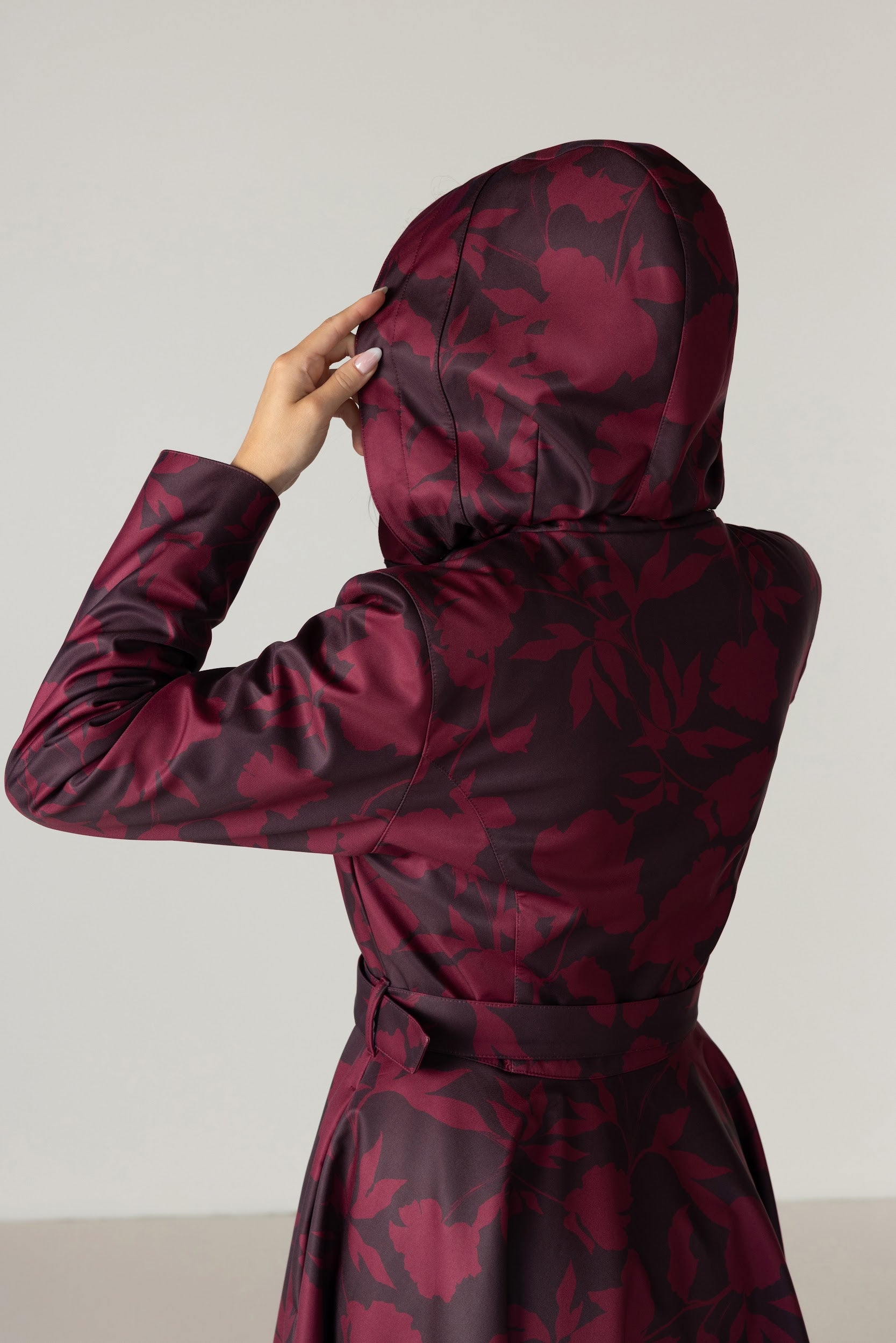 A person shown from the back wearing a hooded long coat with a dark floral pattern in deep burgundy and dark brown by RainSisters, against a plain light gray studio background.
