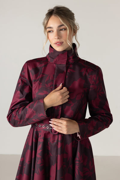 A woman wearing a long-sleeved high-neck coat in a deep burgundy and dark brown abstract pattern by RainSisters, featuring a matching belt with silver grommets, set against a plain light gray studio background.