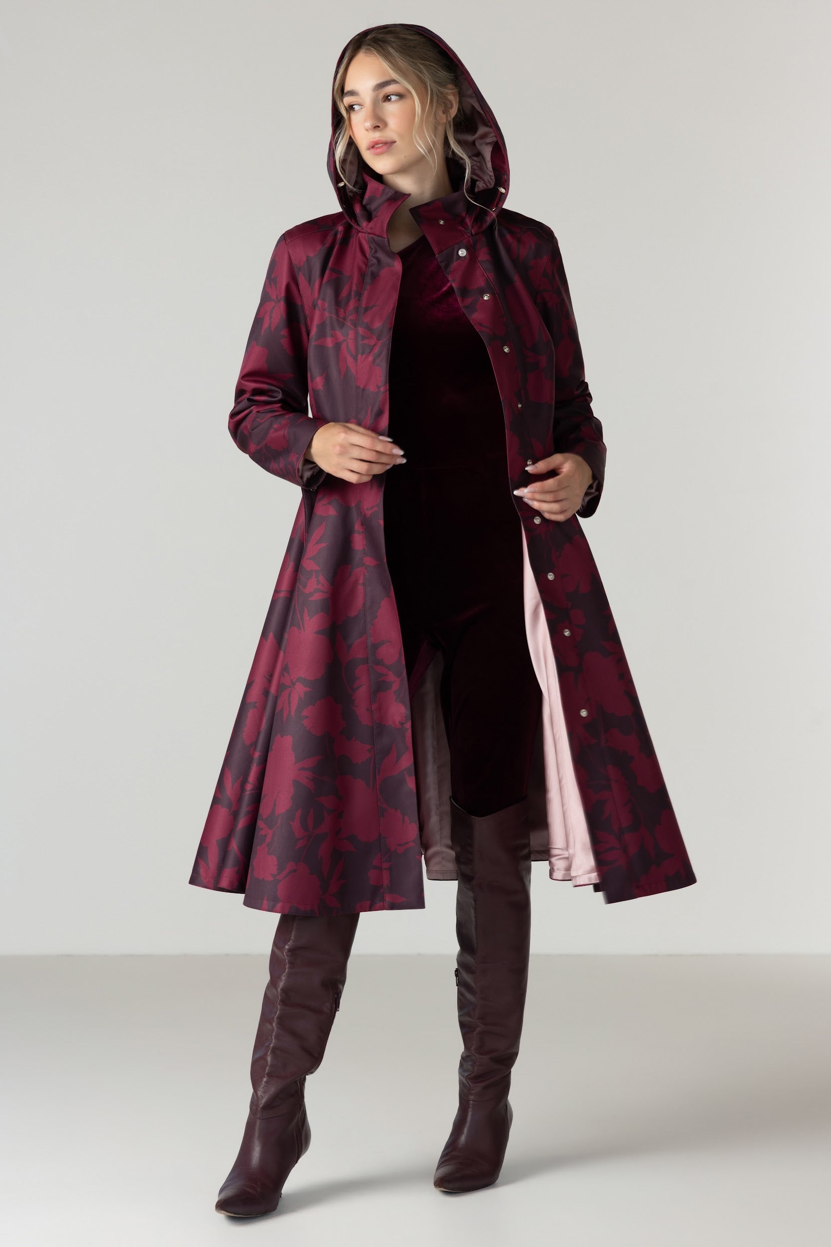 A woman models a long hooded coat with a dark red and black abstract pattern by RainSisters, worn over a dark dress and brown over-the-knee boots against a light gray studio background.