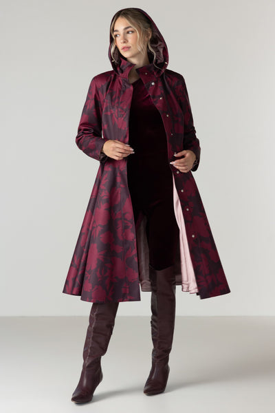 A woman models a long hooded coat with a dark red and black abstract pattern by RainSisters, worn over a dark dress and brown over-the-knee boots against a light gray studio background.