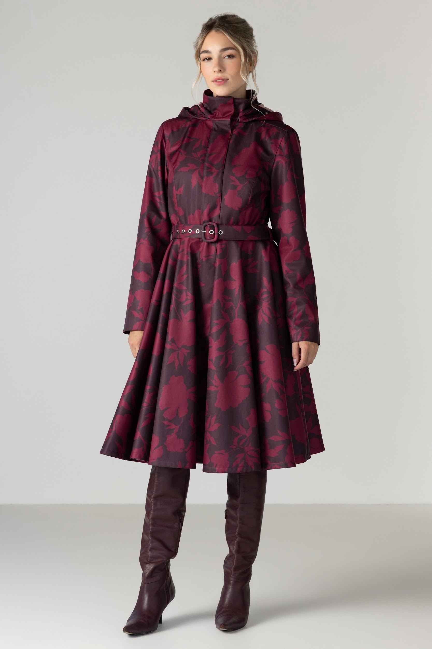 A woman models a long hooded coat with a dark red and black floral jacquard pattern by RainSisters, featuring a belted waist and a flared skirt. She is also wearing dark brown knee-high leather boots against a neutral light gray studio background.