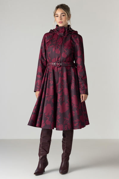 A woman models a long hooded coat with a dark red and black floral jacquard pattern by RainSisters, featuring a belted waist and a flared skirt. She is also wearing dark brown knee-high leather boots against a neutral light gray studio background.