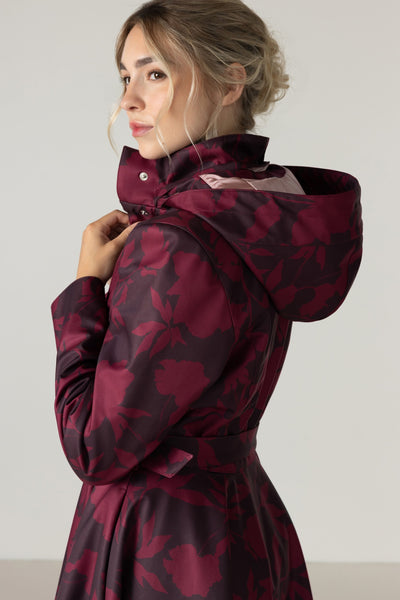 A woman wearing a hooded raincoat with a dark burgundy and dark brown floral pattern by RainSisters, shown from a side-back angle against a plain light gray studio background.