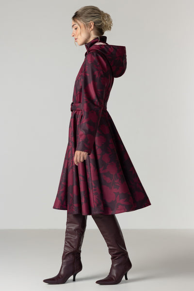 A woman in profile wearing a long hooded coat with a dark red and black floral pattern by RainSisters, paired with dark brown knee-high leather boots, against a light gray studio background.