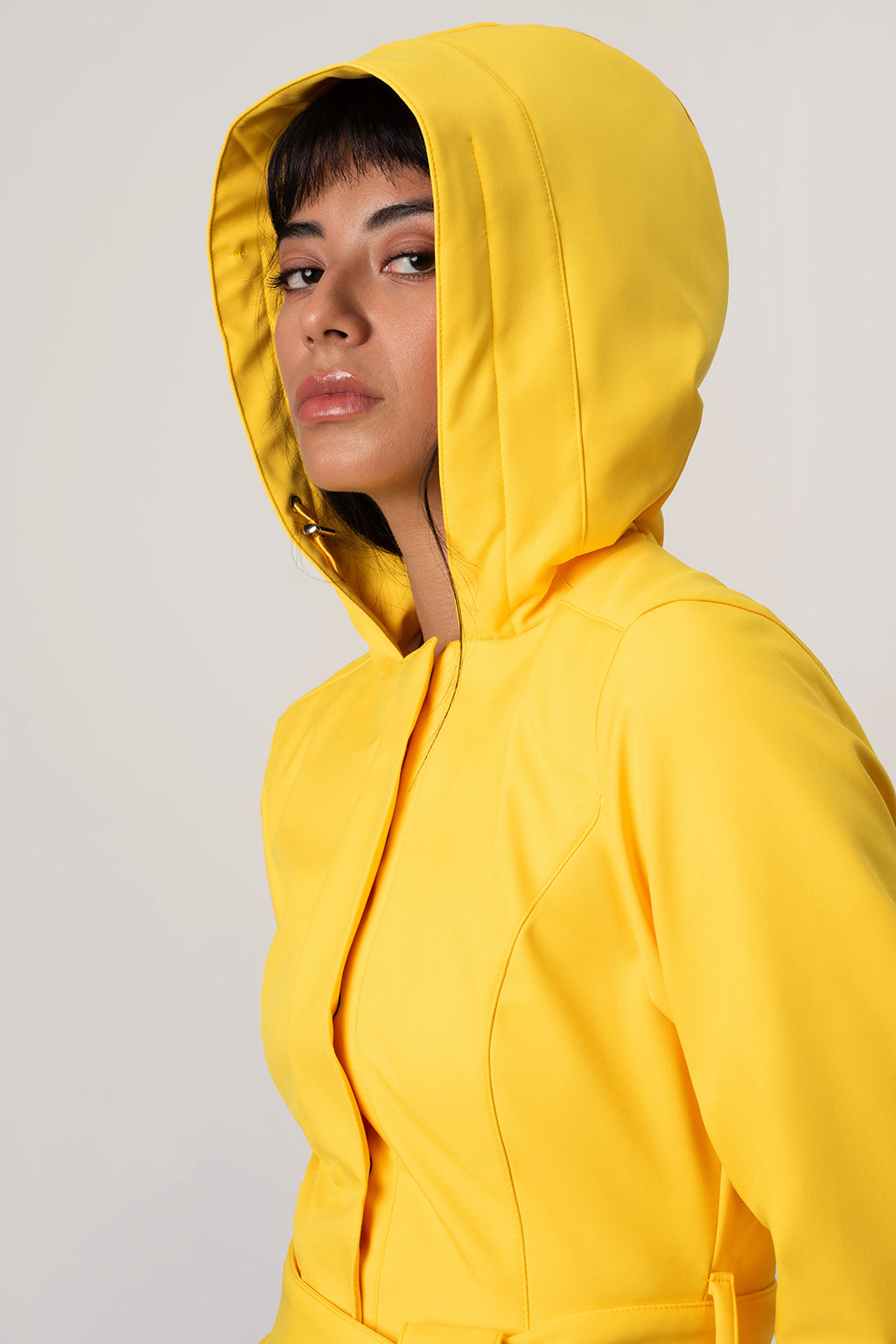 A woman wearing RainSisters bright yellow hooded jacket Yellow Sun, shown in a close-up side profile against a plain light gray studio background.