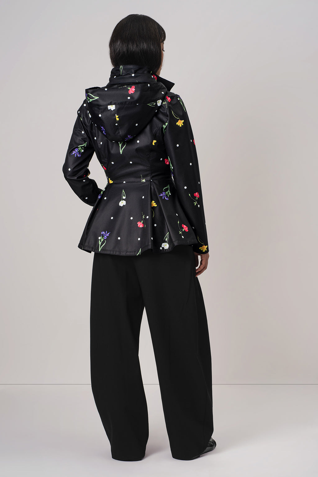 A model shown from the back wearing RainSisters black hooded raincoat Cosmic Meadow with a colorful floral and polka dot print, paired with black wide-leg trousers, against a light gray studio background.